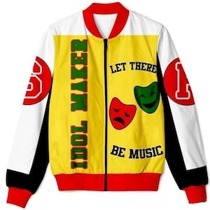 Puede incluir: Una chaqueta bomber amarilla, roja y negra con el texto "Idol Maker" y "Let There Be Music" impreso en la parte delantera. La chaqueta también presenta dos máscaras teatrales, una roja y otra verde.