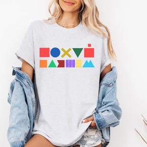 May include: A pale grey t-shirt with a vibrant geometric design. The design features various shapes, including squares, circles, and triangles, in a range of bright colours. The shirt has short sleeves and a relaxed fit.