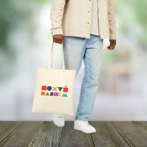 May include: A cream tote bag featuring a vibrant geometric pattern. The design incorporates various shapes like squares and triangles in colours such as red, green, and blue. The bag is held by a person wearing light blue jeans and white trainers.