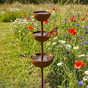 May include: A three-tiered, rustic metal bird bath stands in a vibrant wildflower meadow. The bird bath has three bowl-shaped tiers filled with water. A monarch butterfly rests on the side of the bird bath. Bees are drinking from the bottom tier.