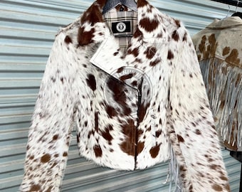 Könnte beinhalten: Kuhfell-Lederjacke mit braun-weißem Fleckenmuster. Die Jacke hat ein fallendes Revers, einen asymmetrischen Reißverschluss und Fransen an den Ärmeln. Die Jacke hängt an einem schwarzen Kleiderbügel.