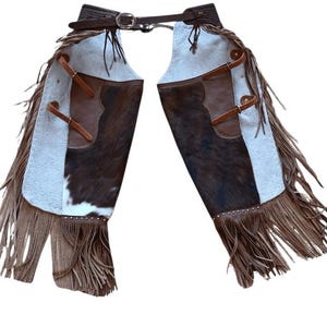 May include: Western-style leather chaps with a brown and white cowhide design. The chaps feature brown leather pockets, straps, and a fringed hem. A dark brown belt with a silver buckle secures the chaps at the waist.