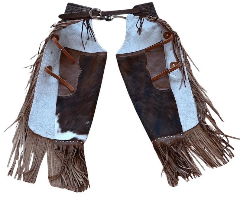 May include: Western-style leather chaps with a brown and white cowhide design. The chaps feature brown leather pockets, straps, and a fringed hem. A dark brown belt with a silver buckle secures the chaps at the waist.