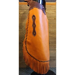 May include: A brown leather chaps with fringe and a brown leather design on the front. The chaps are on a mannequin.