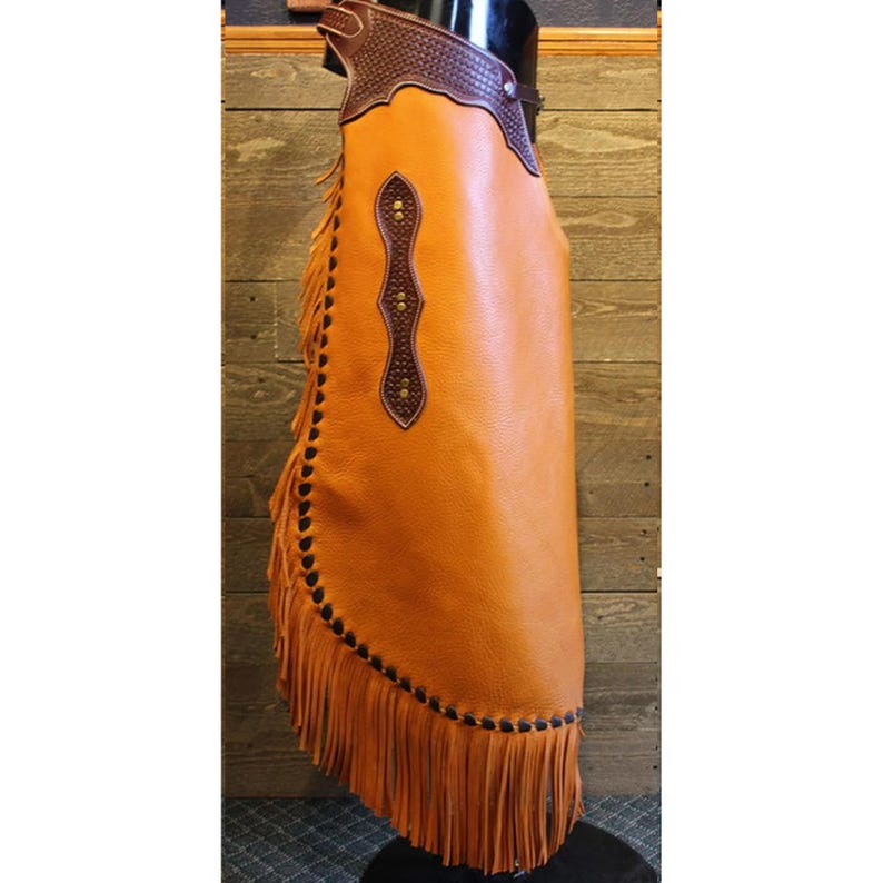May include: A brown leather chaps with fringe and a brown leather design on the front. The chaps are on a mannequin.