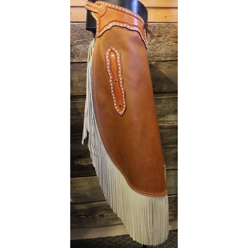 May include: A brown leather chaps with white fringe. The chaps have a decorative stitching detail and a belt loop.