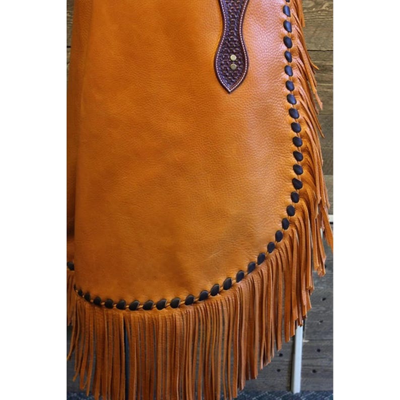 May include: A brown leather chaps with fringe and black stitching. The chaps have a brown leather belt with a decorative buckle.