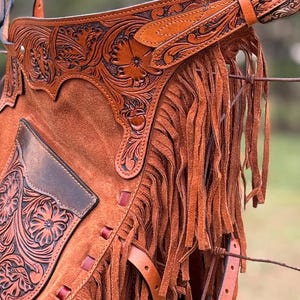 May include: Brown leather chaps with fringe and intricate floral tooling. The chaps have a pocket on the left leg and a belt with a buckle.