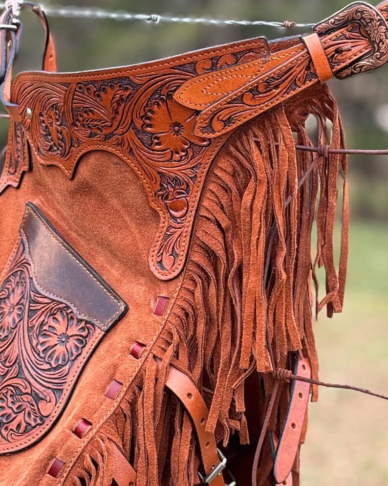 May include: Brown leather chaps with fringe and intricate floral tooling. The chaps have a pocket on the left leg and a belt with a buckle.