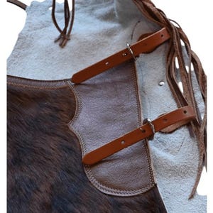 May include: Close-up of a brown and tan leather apron with a fur texture on the lower portion. The apron features brown leather straps with silver buckles and decorative fringe along the side. The apron is designed for protection.