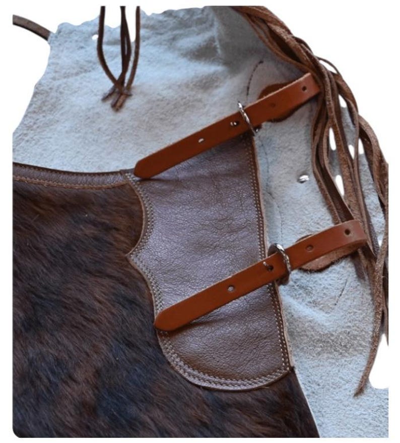 May include: Close-up of a brown and tan leather apron with a fur texture on the lower portion. The apron features brown leather straps with silver buckles and decorative fringe along the side. The apron is designed for protection.