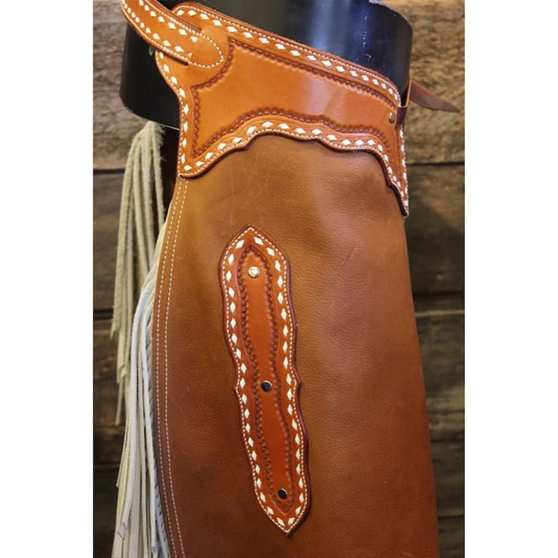 May include: Brown leather chaps with white stitching and fringe. The chaps have a decorative design on the front and back.