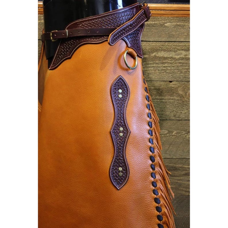 May include: A brown leather chaps with a fringe detail and a brown leather belt with a decorative buckle. The chaps have a brown leather panel with a diamond shape and a decorative stitching detail.