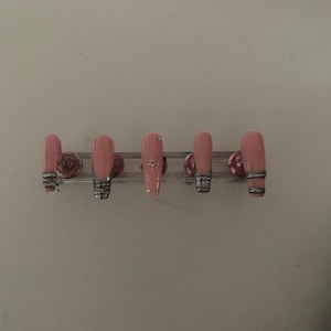 May include: Five pink press-on nails with silver accents. The nails are arranged on a clear plastic display stand.