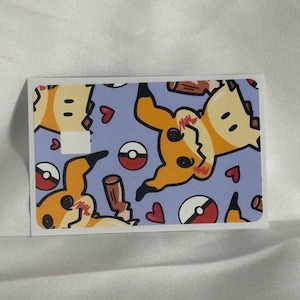 Op de afbeelding: Een rechthoekige sticker met een herhaald patroon van een gele stripfiguur met zwarte accenten, rode harten en rood-witte cirkelvormige ontwerpen op een lichtblauwe achtergrond. De sticker heeft een witte rand.