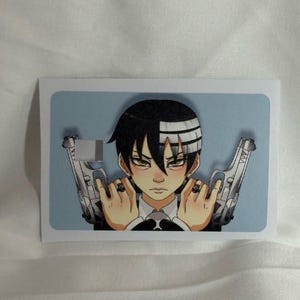 Op de afbeelding: Een rechthoekige sticker met een anime-personage met zwart en wit haar, die twee zilveren pistolen vasthoudt. Het personage heeft een serieuze uitdrukking en draagt een zwart pak met een witte kraag. De achtergrond is lichtblauw.