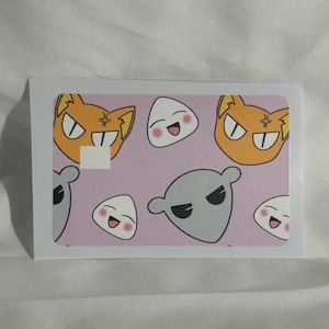 Op de afbeelding: Een rechthoekige sticker met cartoonkarakterontwerpen op een roze achtergrond. De sticker bevat oranje kattengezichten, witte rijstbal-personages en grijze buitenaardse figuren. De sticker is geplaatst tegen een witte achtergrond.