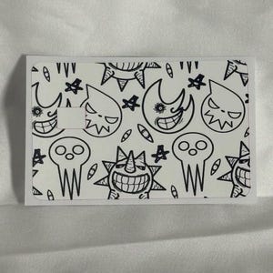 Op de afbeelding: Een witte kaart met een zwart-wit cartoonontwerp. Het ontwerp bevat verschillende lachende gezichten, sterren en andere vormen. De kaart heeft een rechthoekige vorm met een uitsparing aan de linkerkant.