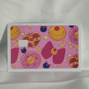 Op de afbeelding: Een rechthoekige sticker met een roze achtergrond met een herhalend patroon van Sailor Moon-elementen. Het ontwerp bevat halve manen, sterren, strikken en hartvormige broches in roze, gele en rode tinten. Een witte rand omlijst de sticker.