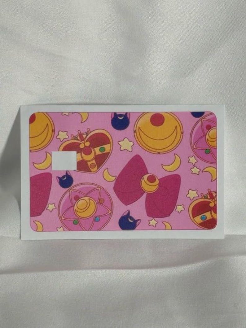Op de afbeelding: Een rechthoekige sticker met een roze achtergrond met een herhalend patroon van Sailor Moon-elementen. Het ontwerp bevat halve manen, sterren, strikken en hartvormige broches in roze, gele en rode tinten. Een witte rand omlijst de sticker.