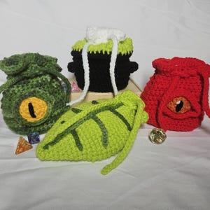 May include: Crocheted dice bags in the shape of fantasy creatures. The bags are green, red, and black, with yellow and orange eye details. A green leaf-shaped pouch and a black cauldron-shaped bag are also present. Dice are scattered around the bags.