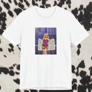 May include: White t-shirt featuring a square image of a person in a pink and black outfit holding a silver cloth. The background is a cityscape. The shirt is on a cow print background.