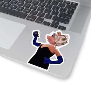 以下が含まれることがあります： ミスピギー、マペットのキャラクターのステッカー。自撮りをしています。黒いドレス、青い手袋、真珠のネックレスを着用しています。彼女のブロンドの髪はアップにまとめられています。ステッカーはラップトップに貼られています。