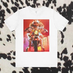 May include: White t-shirt featuring a colorful graphic of a character in a costume riding a motorcycle. The character has a wolf-like mask and is wearing a shiny outfit. The background is a mix of red, orange, and purple hues.