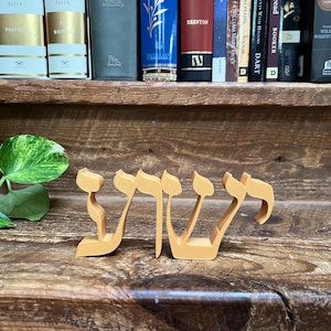 May include: A wooden sign with the Hebrew word "שלום" (shalom) in gold lettering. The sign is sitting on a wooden surface.