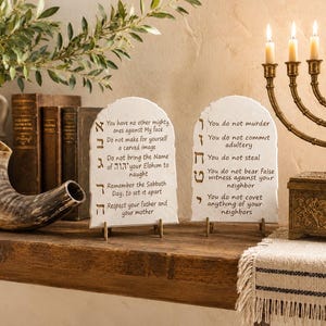 May include: Two white, arched stone tablets featuring black text of the Ten Commandments in English and Hebrew characters. Each tablet is supported by a small metal stand. The text includes commands such as You do not murder, You do not commit adultery, and You do not steal, displayed clearly on the smooth, light-colored surfaces.