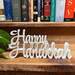 May include: A white, cursive "Happy Hanukkah" sign. The sign is made of a solid material and sits on a wooden surface. The background includes a shelf with books.