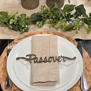 May include: A Passover table setting with a white plate, beige napkin, and a decorative metal word "Passover". The plate rests on a woven placemat, with a fork and knife on either side. Greenery and decorative pots are in the background.