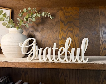 May include: A white, freestanding wooden sign featuring the word "Grateful" in a cursive script font. The letters are connected in a single piece, designed to sit on a flat surface. The sign has a smooth, matte finish and a clean, solid white color.