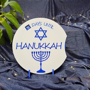 May include: A white Hanukkah countdown sign with blue text and a blue Star of David and menorah design. The sign reads "30 Days Until Hanukkah". The sign is propped up by two blue stands.