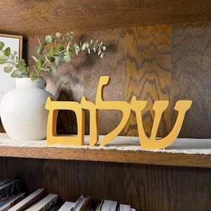 May include: A mustard yellow freestanding wooden sign displaying the Hebrew word Shalom in a bold, sans-serif font. The letters are connected in a single piece, featuring a smooth matte finish and a clean, modern aesthetic for shelf display.