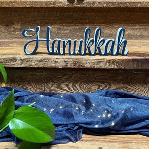 May include: A blue and white wooden sign that reads "Hanukkah" in a cursive font. The sign is placed on a wooden surface with a blue fabric and string lights in the background. Green leaves are in the foreground.