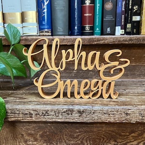 May include: A wooden sign with the words "Alpha & Omega" in a flowing script. The sign is a warm, golden color and sits on a wooden surface. The background includes a shelf with books and greenery.