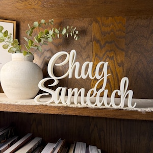 May include: A white, three-dimensional script sign featuring the text Chag Sameach in a cursive font. The sign is positioned next to a white, textured ceramic vase containing green eucalyptus stems, creating a decorative display.