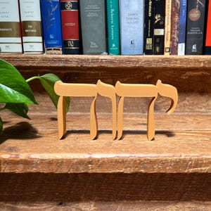 May include: Wooden Chai symbol in a light brown color, standing on a wooden surface. The Chai symbol is a Hebrew word meaning "life". Books are visible in the background.
