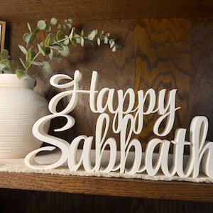 May include: A white, three-dimensional script sign featuring the words "Happy Sabbath" in a cursive font. The letters are connected and stand upright on a flat surface, displaying a smooth, matte finish with clean, rounded edges throughout the text.
