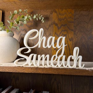 May include: A white, three-dimensional script sign featuring the text "Chag Sameach" in a cursive font. The letters are connected and stand upright on a flat surface, displaying a clean, matte finish with smooth, rounded edges throughout.