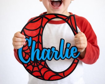 Könnte beinhalten: Ein Holzausschnitt in Form des Buchstabens "C" mit einem roten Spinnennetz-Design und dem Namen "Charlie" in blauer Schrift. Der Ausschnitt wird von einem Kind gehalten, das ein rot-weißes Hemd trägt.