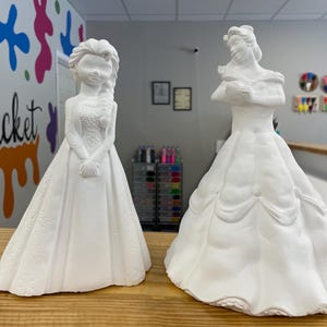 Könnte beinhalten: Zwei weiße Keramikfiguren von Prinzessinnen, eine mit einem langen, fließenden Kleid und die andere mit einem enganliegenden Kleid. Beide Figuren stehen auf einer Holzoberfläche.