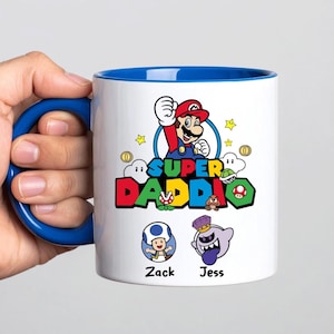 May include: White ceramic mug with a blue interior and handle. The mug features a colourful graphic of Mario with the text "SUPER DADDIO" and character illustrations. The names "Zack" and "Jess" are also printed on the mug.