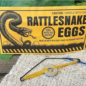 May include: A yellow and black sign with the words "Rattlesnake Eggs" and a graphic of a snake. The sign also includes the text "Caution: Handle with Care" and "100% Reptile Egg Certified". A small slingshot is in front of the sign.