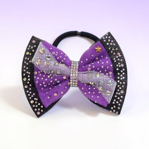 May include: A purple hair bow with a black border, adorned with rhinestones and a gold star. The bow features a central rhinestone band and is attached to a black elastic hair tie. The bow is a decorative hair accessory.