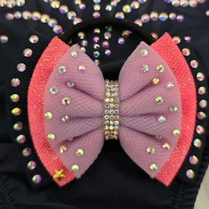May include: A close-up of a hair bow with a layered design. The bow features a light purple mesh center, framed by a coral and black layer, and adorned with iridescent rhinestones. A gold star is visible on the black layer.