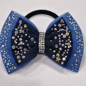 May include: A blue hair bow with a black elastic band. The bow features a layered design with a darker blue center and lighter blue edges, embellished with sparkling rhinestones and a small gold star.
