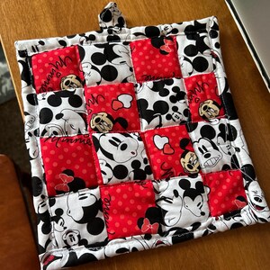 May include: A red and white quilted hot pad featuring a Mickey Mouse and Minnie Mouse design. The hot pad has a loop handle and is made of cotton fabric.