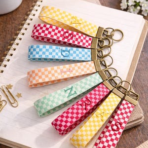 Pode incluir: Uma coleção de chaveiros coloridos xadrez com ferragens douradas. Cada chaveiro apresenta uma letra diferente bordada na alça. Os chaveiros estão dispostos em um caderno, com uma xícara de café ao fundo.